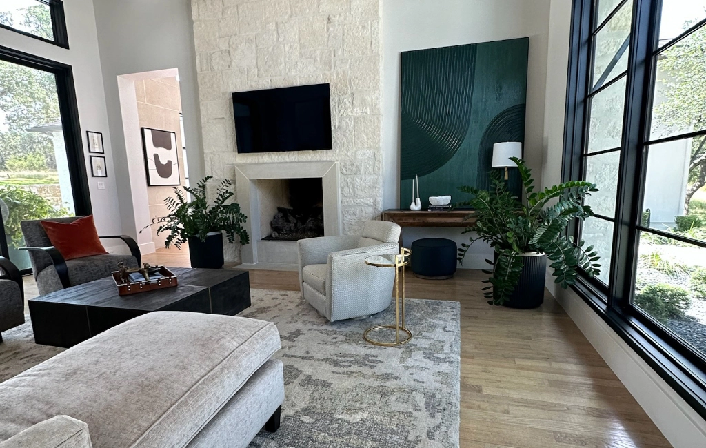 A modern living room with a large window, stone fireplace, wall-mounted TV, and contemporary furniture. Houseplants and abstract artwork on the walls enhance the minimalist decor.