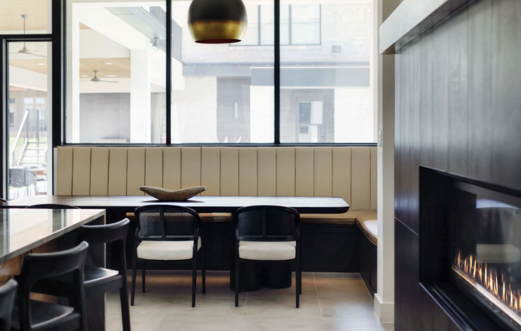 Modern dining area with a long bench, four black and white chairs, a black table, and large windows letting in natural light. A gold and black pendant light hangs above the table.