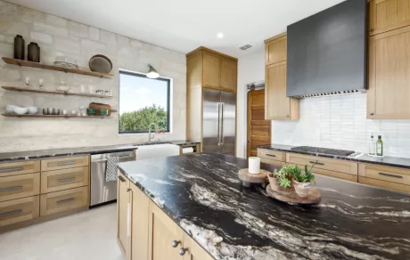 Modern kitchen featuring wooden cabinets, marble countertops, open shelving, a large island with a sink, and stainless steel appliances. A window provides natural light.