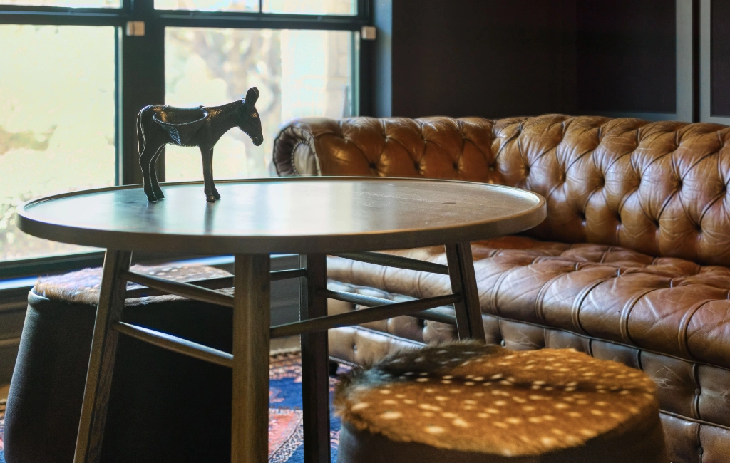 A room with a leather tufted sofa, a round wooden table with a small donkey figurine, and two stools with animal print upholstery. Natural light filters through the window.