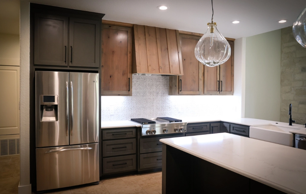 Modern kitchen with stainless steel refrigerator, oven, and brown cabinetry. Features white countertops, a farmhouse sink, and a large glass pendant light.