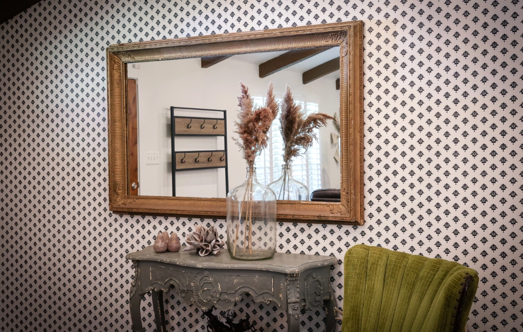 A large mirror with a wooden frame hangs on a patterned wall. Below it is a decorative table with a glass vase holding dried flowers and a green chair beside it.