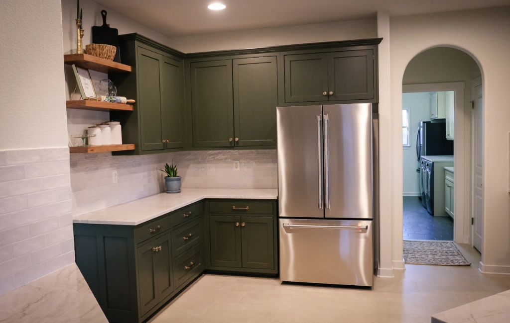 A kitchen with green cabinets, stainless steel fridge, white countertops, and open shelves. An arched doorway leads to another room.