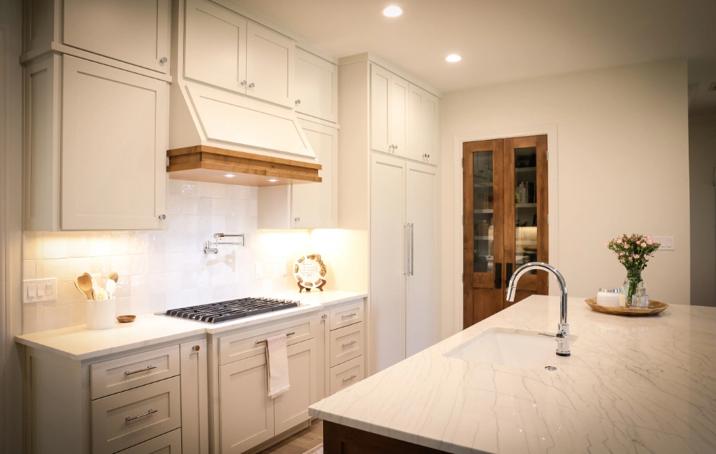 A modern kitchen with white cabinets, a marble island with a sink, a gas cooktop with a wooden hood, and a vase of flowers on the countertop.