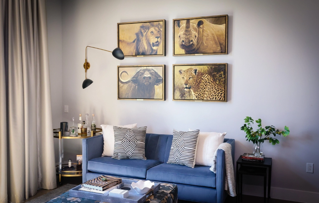 A living room with a blue sofa, two patterned throw pillows, two wall lamps, and four framed wildlife photos above. A side table holds bottles and glasses, and a small plant is beside the sofa.