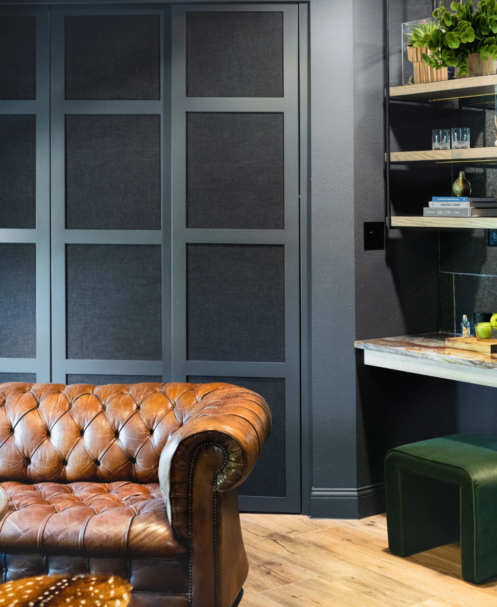 A cozy room features a brown leather tufted sofa, dark paneled walls, wooden flooring, and a small desk area with shelves decorated with plants and books.