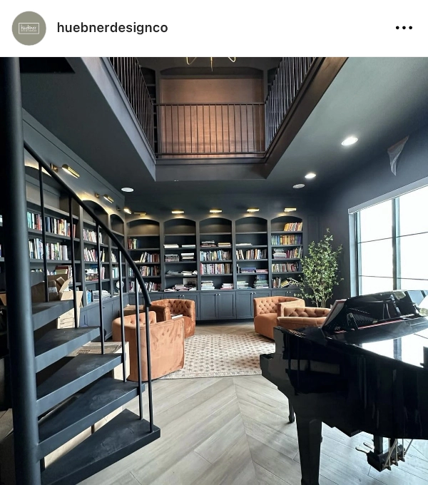 A modern library with black shelving and a spiral staircase. Orange chairs, a grand piano, and a large window add to the room's design. A second-floor balcony overlooks the space.