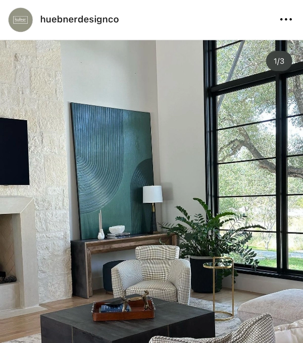 Modern living area featuring a green abstract painting, large windows, white armchairs, a stone fireplace, a potted plant, and a small table with a lamp.