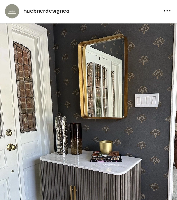 A modern entryway features a white door with glass panels, a patterned wallpaper, a console with a marble top, decorative vases, books, and a gold-framed mirror above.