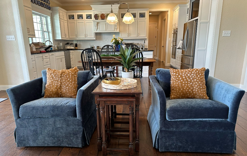 Spacious kitchen with white cabinetry and stainless steel appliances, overlooking a sitting area with two blue armchairs, a wooden side table, and a dining table with chairs. A potted plant decorates the table.