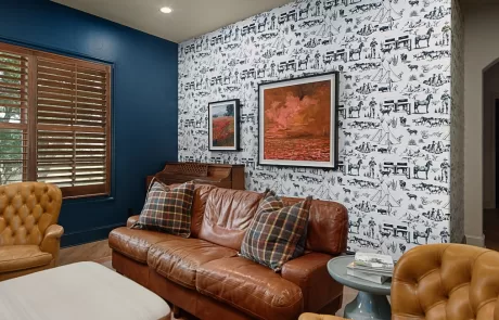 A living room with a brown leather sofa, patterned wallpaper, framed artwork, wooden shutters, and a small round side table. The walls are partially blue and accented with designs.