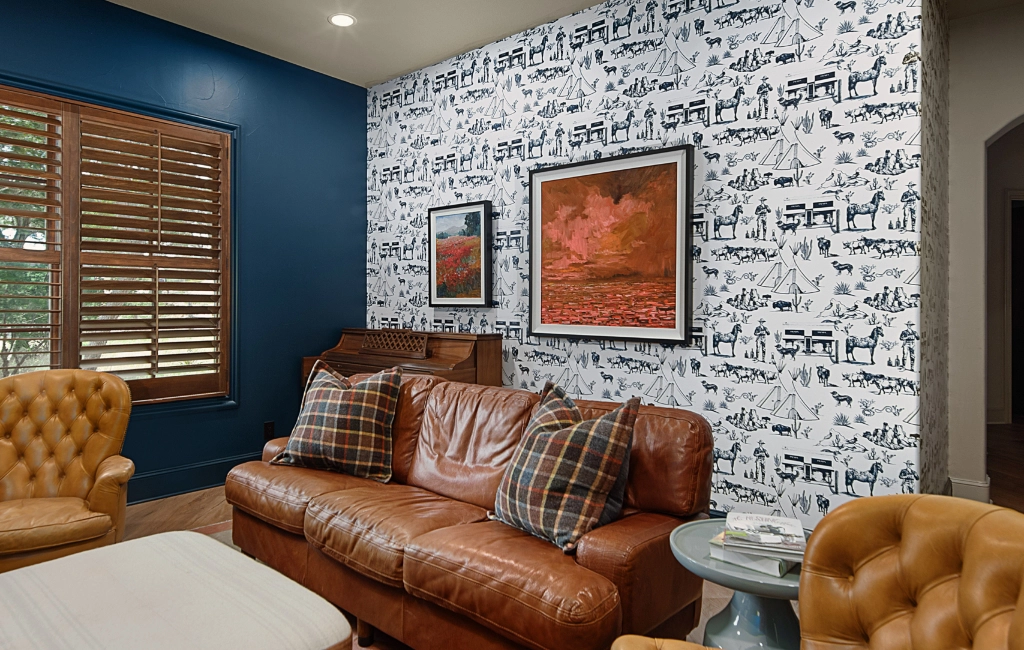 A living room with a brown leather sofa, patterned wallpaper, framed artwork, wooden shutters, and a small round side table. The walls are partially blue and accented with designs.