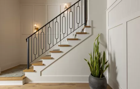 A modern staircase with wooden steps and a black metal railing, adjacent to a white paneled wall with a potted plant at the base.