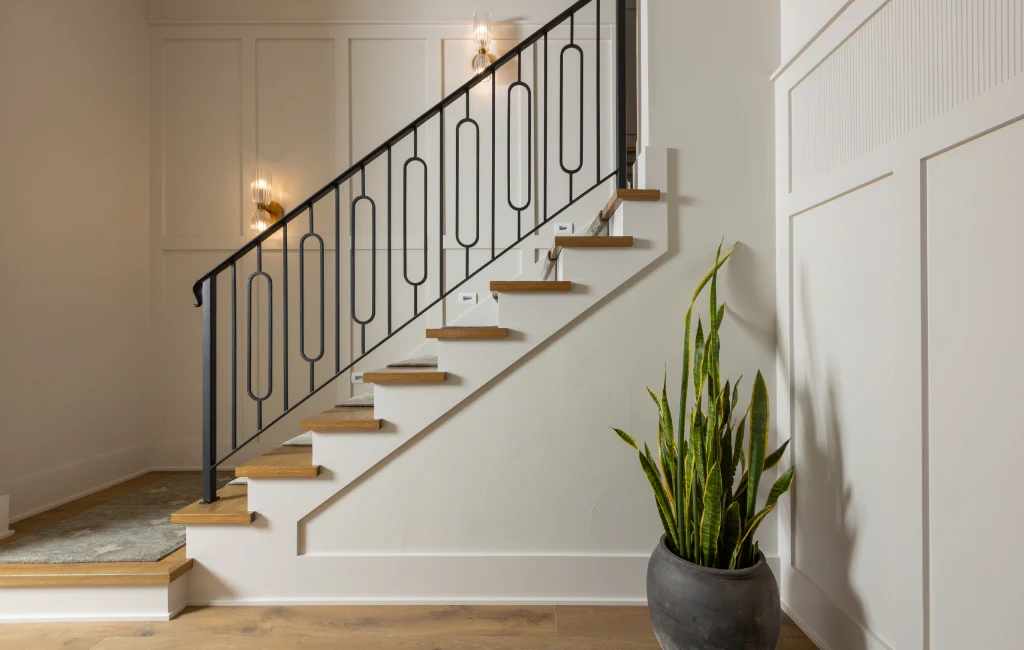 A modern staircase with wooden steps and a black metal railing, adjacent to a white paneled wall with a potted plant at the base.