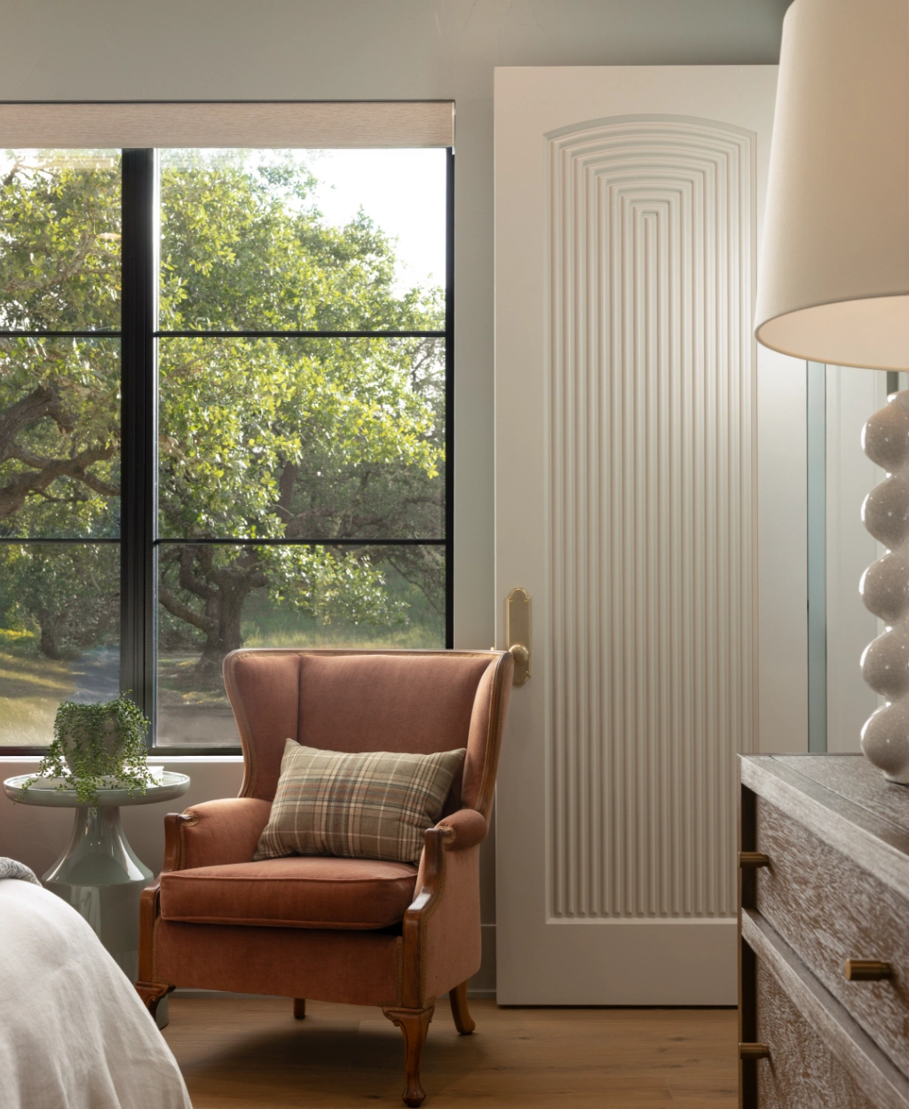 A pink upholstered armchair with a plaid pillow sits by a window next to a small round table and a white textured door in a bright, modern room.