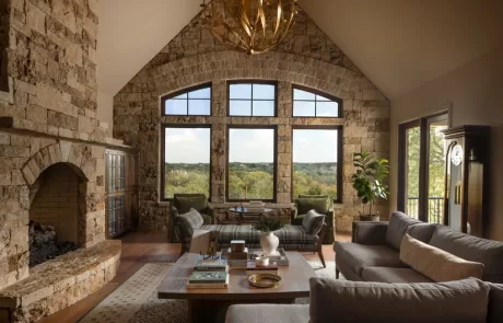 Rustic living room with vaulted ceiling, stone fireplace, large arched windows, and modern furniture overlooking Hill Country landscape