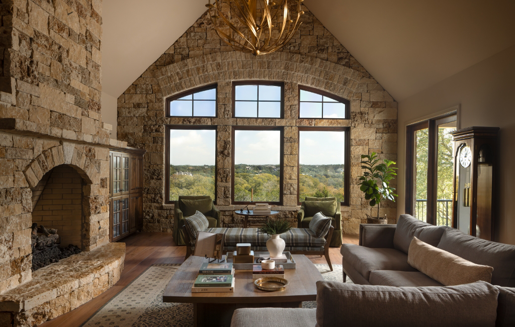 Rustic living room with vaulted ceiling, stone fireplace, large arched windows, and modern furniture overlooking Hill Country landscape